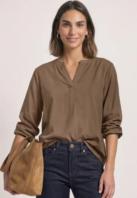 Frau trägt braune Bluse und Jeans, hält braune Wildledertasche, lächelnd vor hellem Hintergrund.