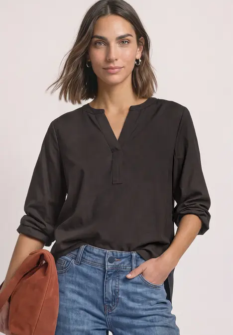 Frau in schwarzer Bluse mit V-Ausschnitt, hochgekrempelten Ärmeln und Jeans, die eine braune Handtasche hält.