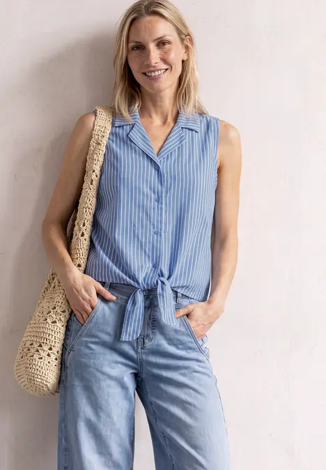 Eine lächelnde Frau in ärmelloser, hellblauer Bluse mit Knoten, Jeans und Strohtasche lehnt an weißer Wand.