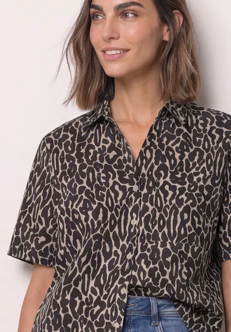 Eine lächelnde Frau trägt eine kurzärmlige Bluse mit braun-schwarzem Leopardenmuster und offenem Kragen.