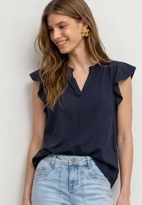 Lächelnde Frau trägt dunkelblaue Rüschenbluse und helle Jeans.