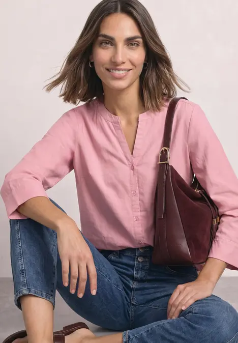 Frau sitzt lächelnd, trägt rosa Bluse, blaue Jeans und hält eine Handtasche.