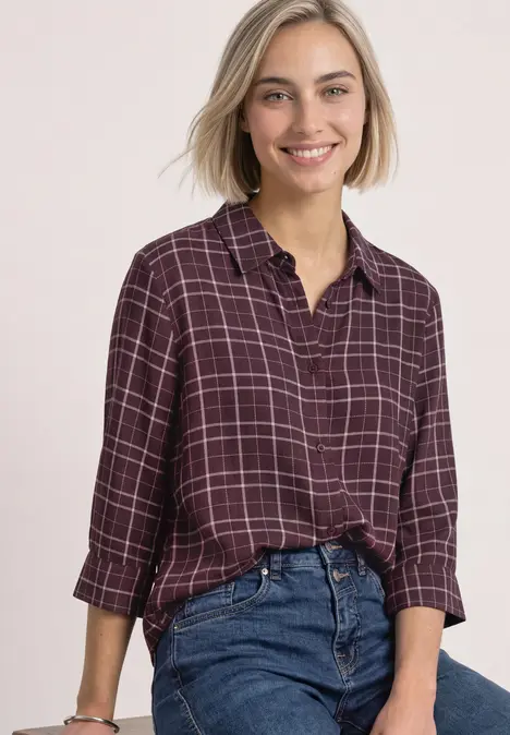Eine lächelnde Frau in einem karrierten weinroten Hemd, die locker eine Jeans trägt vor hellem Hintergrund.