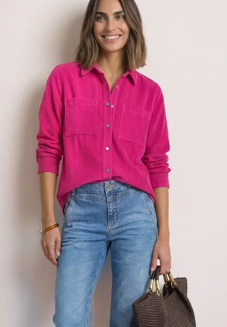 Frau in pinkem Hemd und blauen Jeans lächelt und hält eine braune Handtasche.