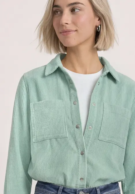 Frau mit kurzem blonden Haar trägt hellgrünes Kordhemd über weißem T-Shirt.