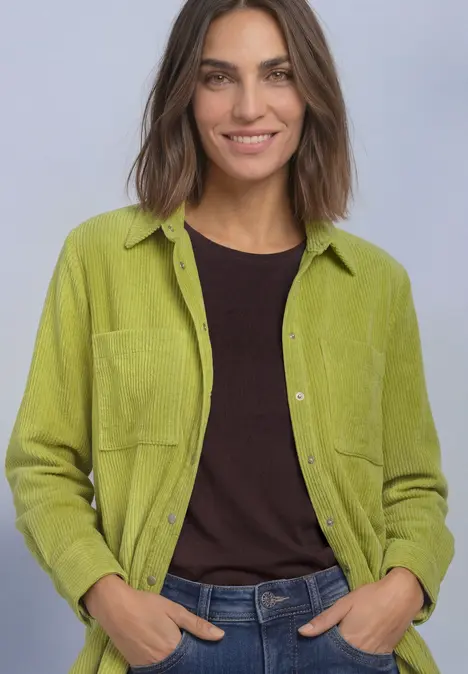 Frau mit grüner Hemdjacke, schwarzem Shirt und Jeans, lächelt in die Kamera.