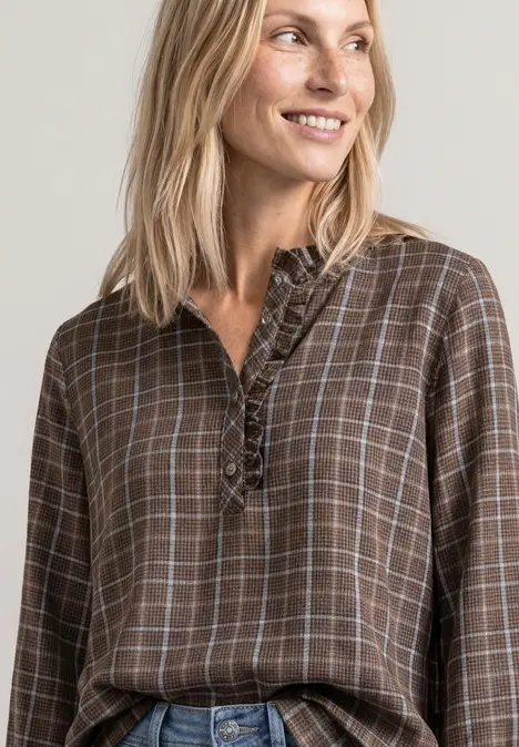 Blonde Frau lächelt seitlich, trägt braun-kariertes Blusenshirt mit Rüschen vor neutralem Hintergrund.
