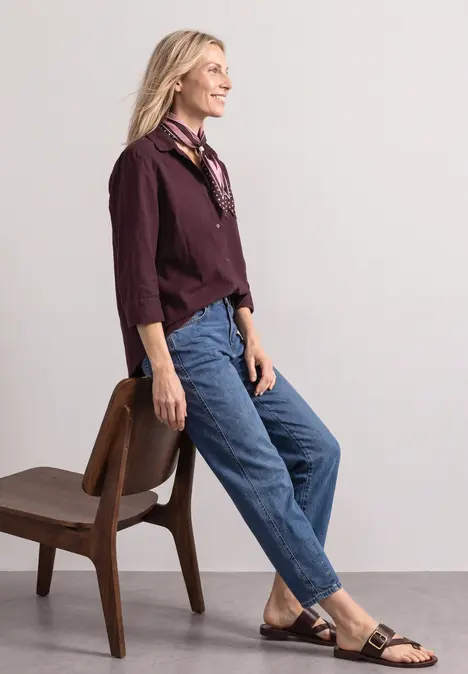 Eine Frau lehnt an einem Stuhl, trägt Jeans, bordeauxfarbenes Hemd, schwarze Sandalen und lächelt zur Seite.