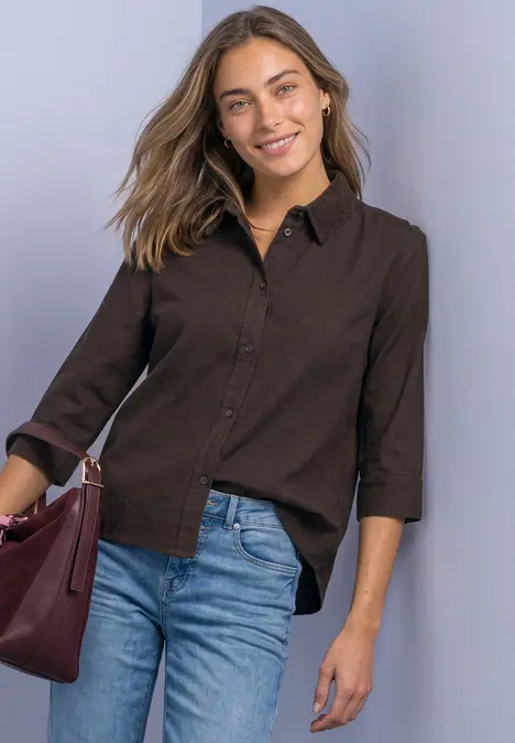 Frau in brauner Bluse und Jeans hält eine rote Handtasche und lächelt.