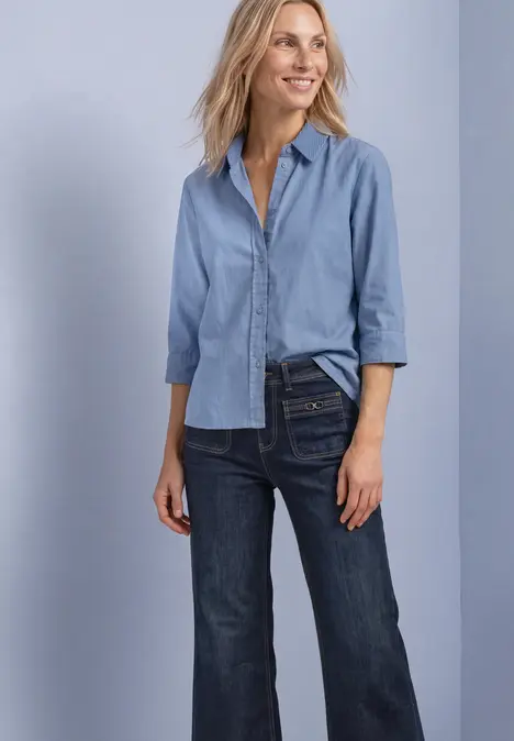 Dame trägt hellblaue Bluse und dunkle Jeans, lächelt vor hellem Hintergrund.
