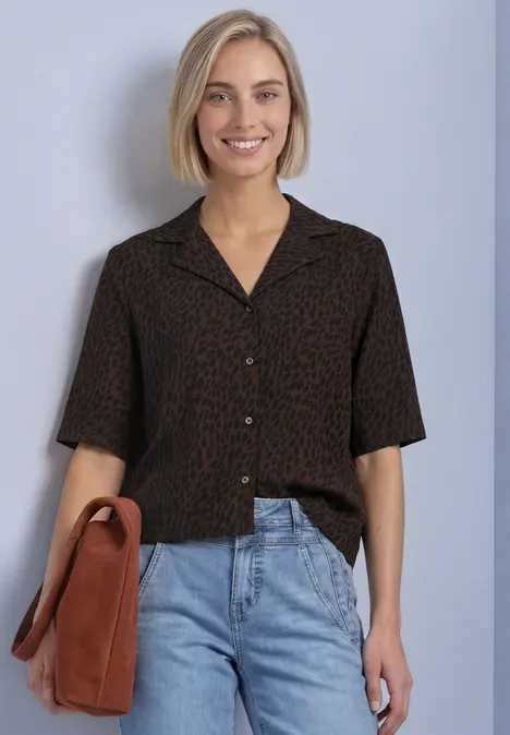 Junge Frau in kurzärmeligem, braun-schwarzem Print-Hemd, Jeans und Umhängetasche, lächelnd