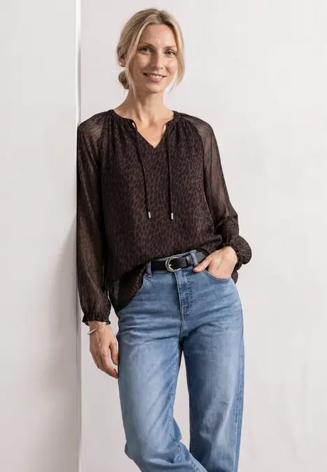 Frau mit schwarzer gepunkteter Bluse und blauen Jeans lächelt vor weißem Hintergrund.