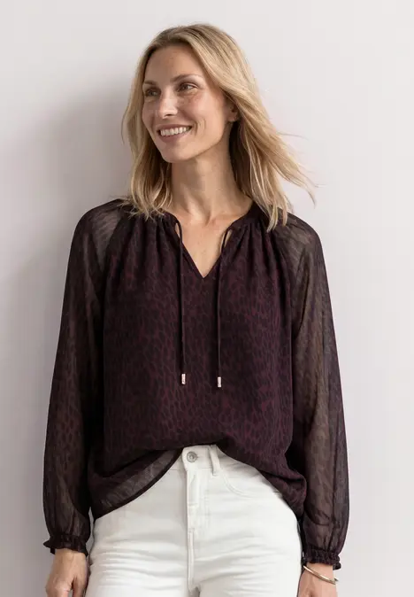 Eine lächelnde Frau trägt eine dunkle, gemusterte Bluse mit V-Ausschnitt und weiße Jeans.