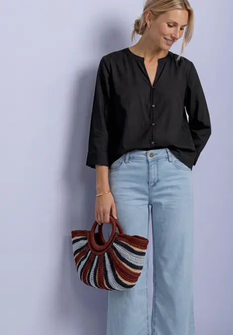 Frau in schwarzer Bluse und hellblauen Jeans hält bunte gestreifte Tasche vor lila Hintergrund.