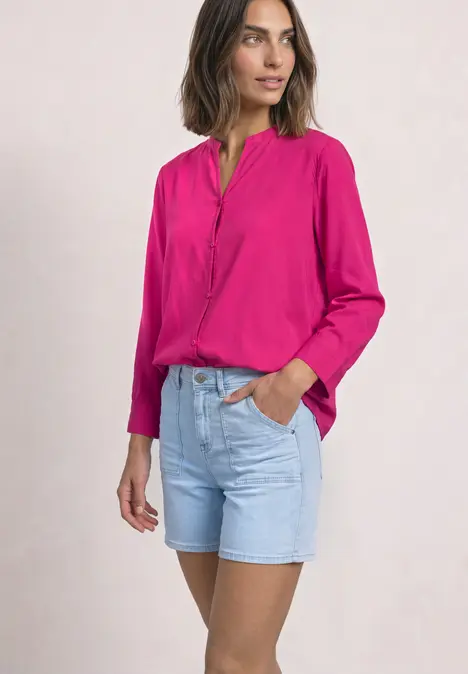 Frau trägt pinke Bluse und hellblaue Jeans-Shorts, steht lässig mit einer Hand in der Tasche.