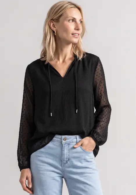 Blonde Frau in schwarzer Chiffon-Bluse mit V-Ausschnitt und hellen Jeans, Blick seitlich, Hand in Tasche.