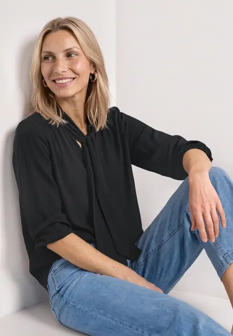 Eine Person sitzt mit schwarzer Bluse und blauen Jeans an einer hellen Wand.