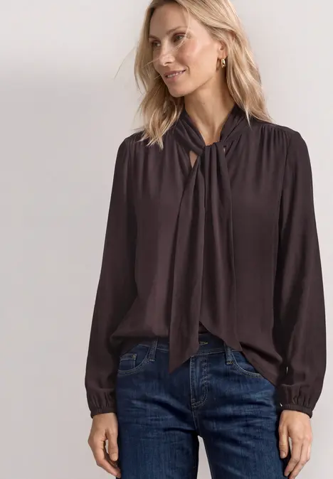 Frau trägt dunkelbraune Bluse mit Knoten am Kragen und Jeans.