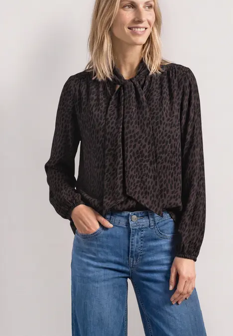 Eine Frau trägt eine schwarze, gemusterte Bluse mit Schleife am Hals und blaue Jeans.