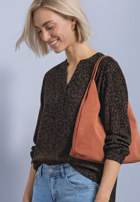 Eine Frau in gemustertem Pullover, Jeans und braun-orangener Tasche lächelt und blickt nach unten.