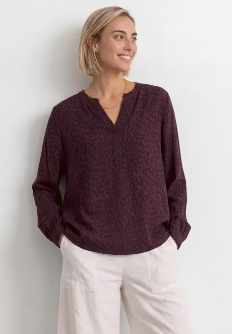 Eine lächelnde Frau mit kurzen blonden Haaren trägt eine weinrote gemusterte Bluse und helle Hose.