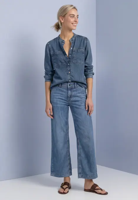 Eine lächelnde Frau trägt Jeanshemd, weite Jeans und braune Sandalen vor hellgrauer Wand.