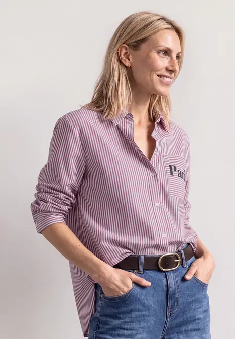 Femme en chemise rose à rayures et jean, les mains dans les poches, regard détourné.