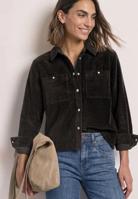 Vrouw glimlacht in zwart corduroy overhemd en blauwe jeans met een beige tas in de hand.