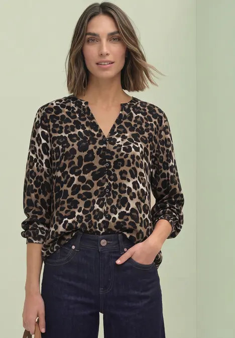 Frau mit schulterlangem Haar trägt Leopardenbluse und hohe dunkelblaue Jeans vor hellem grünen Hintergrund.