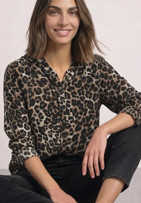 Uśmiechnięta kobieta z brązowymi włosami ma na sobie panterkową bluzkę i czarne spodnie na jasnym tle.