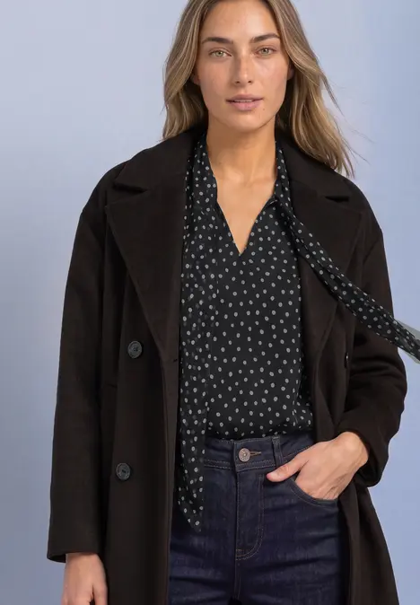 Frau in schwarzem Mantel, gepunkteter Bluse und Jeans vor hellem Hintergrund.