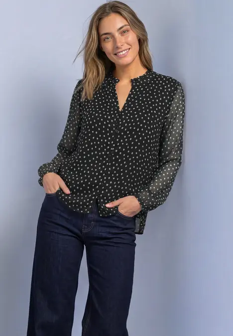 Eine Frau steht lächelnd, trägt eine schwarze, gepunktete Bluse und dunkle Jeans.