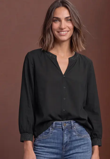 Eine lächelnde Frau in schwarzem Blusenshirt und Jeans vor braunem Hintergrund.