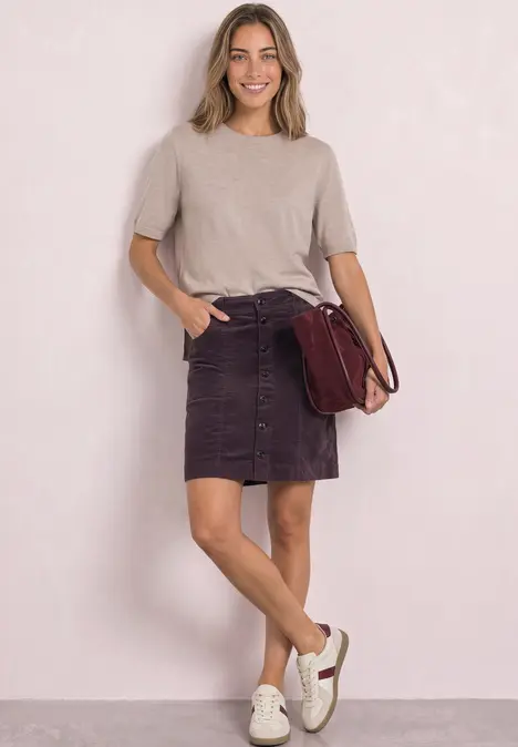 Junge Frau mit beigem Oberteil, violettem Knopfrock, weißen Sneakern und roter Schultertasche, lächelnd stehend.