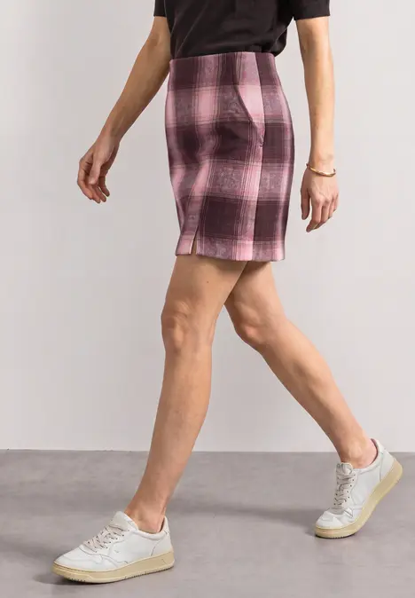 Femme en haut noir, jupe courte rose à carreaux et baskets blanches marchant.