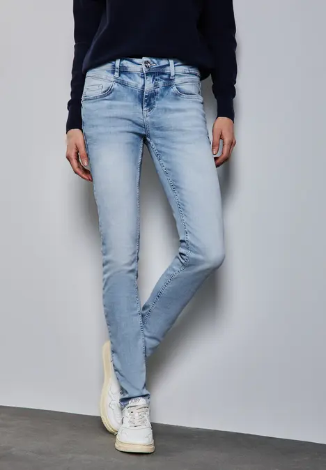Person trägt eine helle Jeans und weiße Sneakers, steht vor einer grauen Wand.