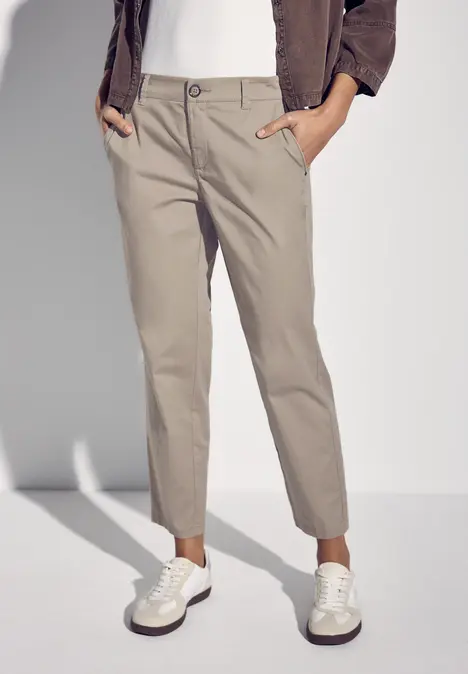 Beige Chinohose, weiße Sneaker, braune Jacke, Hände in den Taschen.