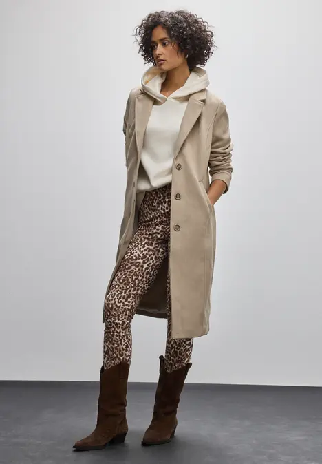 Frau trägt Leo-Leggings, beigen Mantel und weiße Sneaker.
