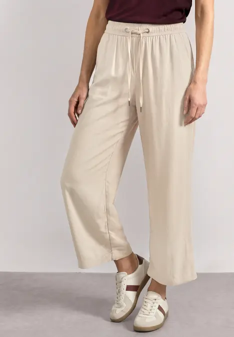 Beine in beigen, weiten 7/8-Hosen und weißen Sneakern mit beige-hellen Details vor neutralem Hintergrund.