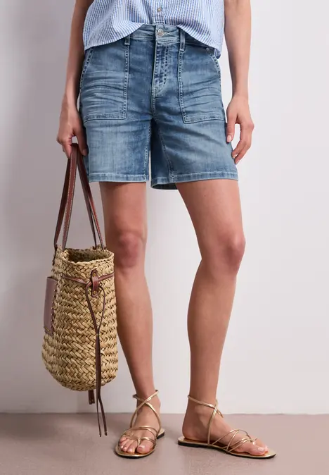 Beine in Jeans-Shorts, geflochtene Strohtasche und goldene Riemchensandalen.