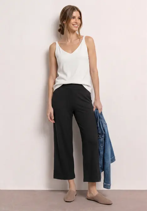 Frau in weißem Top, schwarzen Culottes und taupe Mules, mit Jeansjacke in der Hand.