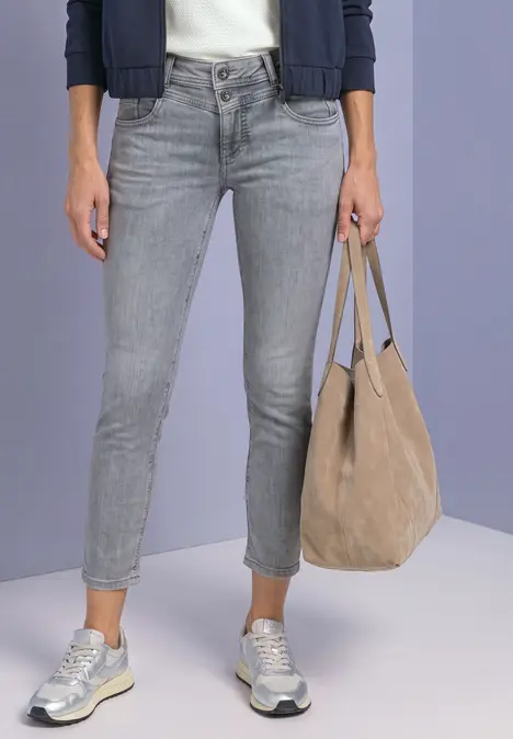 Eine Person trägt graue Skinny-Jeans, silberne Sneaker und hält eine beige Veloursleder-Tasche.
