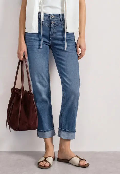 Frau trägt hochgekrempelte blaue Jeans, weiße Weste, Sandalen und braune Wildledertasche.