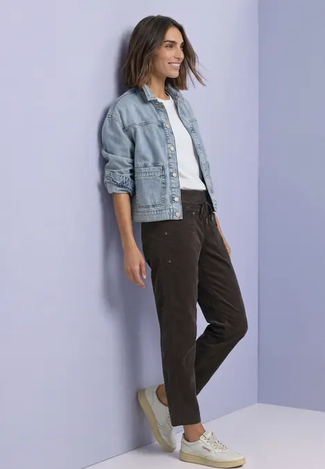 Eine lächelnde Frau in Jeansjacke, weißem Shirt und Cordhosen lehnt an einer pastellfarbenen Wand.