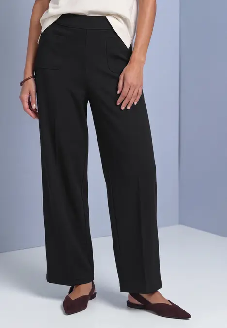 Eine Person trägt schwarze, weite Hosen und spitze, bordeauxfarbene Ballerinas.