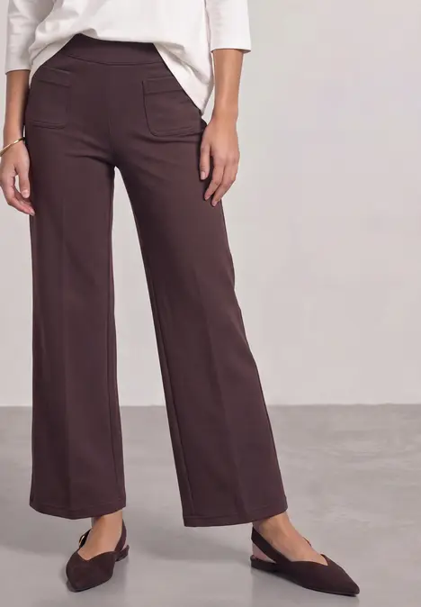 Person trägt weite, braune Stoffhose, weiße Bluse und spitze Ballerinas.