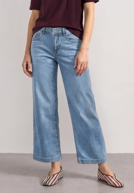 Eine Person trägt helle, weite Jeans mit weitem Bein und gestreiften, spitzen Flats vor neutralem Hintergrund.