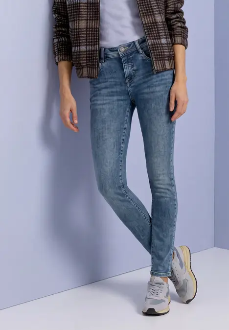 Person mit schmalen blauen Jeans, kariertem Blazer und weißen Sneakers vor einfarbigem Hintergrund.
