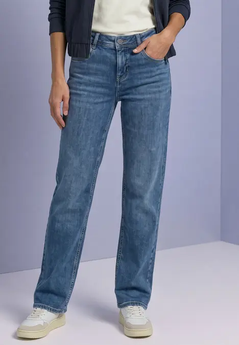 Eine Person trägt gerade geschnittene blaue Jeans und weiße Sneaker vor neutralem Hintergrund.