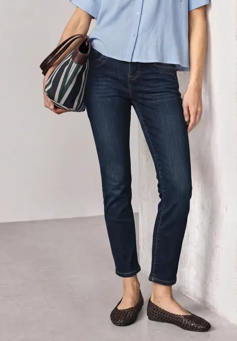 Person in dunklen Jeans, hellblauer Bluse und gemustertem Umhängetasche, steht entspannt an Wand